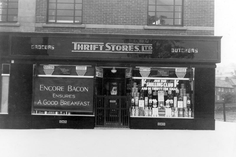 Shop window displays of Thrift Stores, grocer and butchers shop in September 1936. A message on the window blind reads 'Encore Bacon Ensures a Good Breakfast'. The right hand window has an invitation to 'Join Our Shilling Club, and be thrifty for Xmas'.