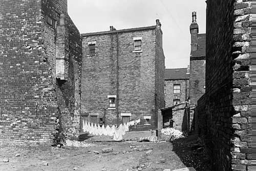 This view looks across an area of waste ground to the rear of numbers 68 and 70 Glover Street, which are in the centre. To the left are the backs of numbers 8 and 10 Gerrans Street, which appear to be in the process of demolition, while on the right is an outside toilet. Pictured in July 1958.