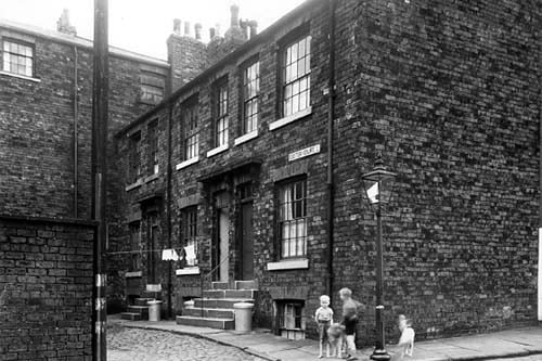 Looking from Exeter Street to Exeter Court where three houses can be seen. On the left is the back wall of 4/6 Exeter Place, then moving right, numbers 3, 4 and 5 Exeter Court. Two boys are on the corner, next to the street light. These and the surrounding streets were demolished to provide part of the site for the Inner Ring Road, being between Woodhouse Lane, Clay Pit Lane and Carlton Hill Barracks. Pictured in August 1959.