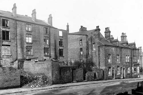 A view of derelict houses, which front Northfield Terrace at the other side, viewed from Albert Grove in October 1950. The four storey houses on the left have broken and bricked up windows. The houses on the right are occupied. Tiger's Grocers is at the junction with Trafalgar Terrace on the far right, at numbers 53 - 55 Albert Grove. The odd-numbered side of Albert Grove began with number 1, off camera, left, at the junction with Lovell Road. The enclosing garden walls with doors lead onto the cobbled street. A small boy is sitting in the doorway of the house above the shop.