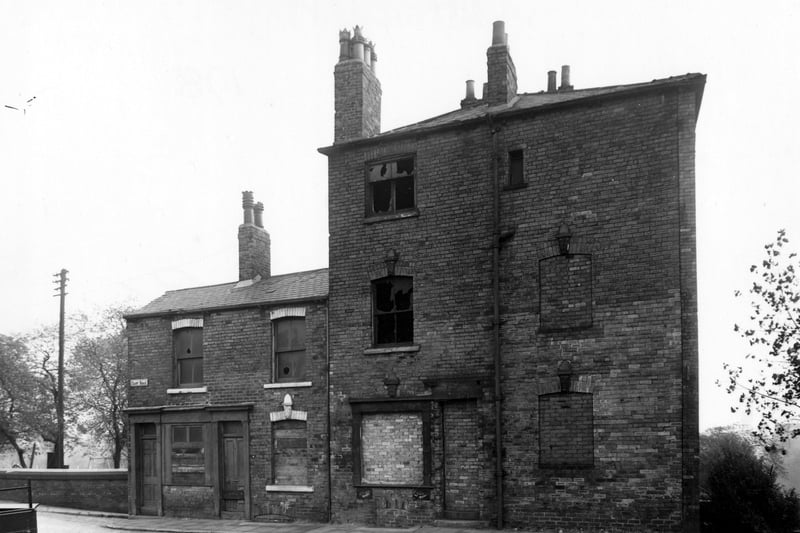 Two abandoned brick buildings containing three dwellings on Camp Road in October 1955. They have some ornate chimney pots and window heads. Just visible in the background is a playground. The road in the foreground is cobbled.